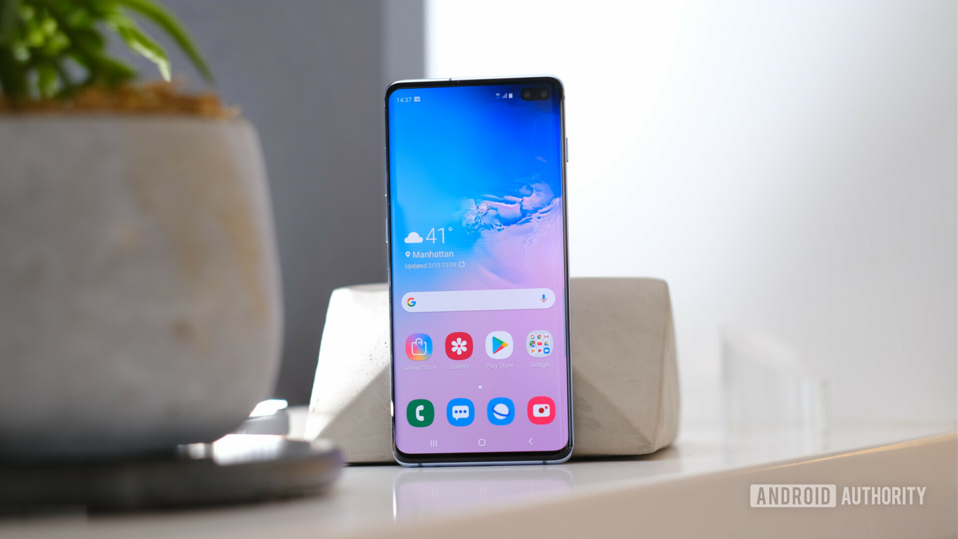 Frontside photo of the new Samsung Galaxy S10 Plus standing upright on a table with a screen turned on.