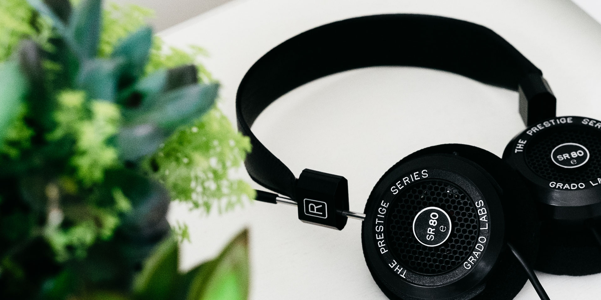 Grado SR80e headphones product lifestyle image from Grado. Headphones are on a white table with a plant in the left side of the frame.