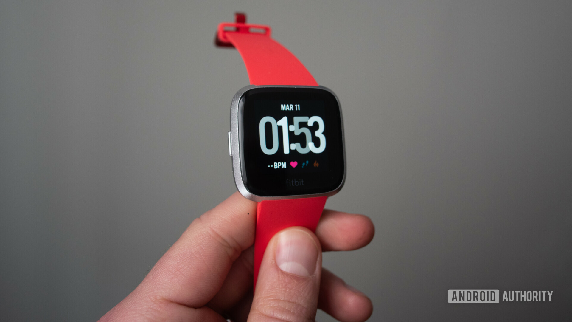 Front side of the Fitbit Versa Lite with a red strap held in hand.