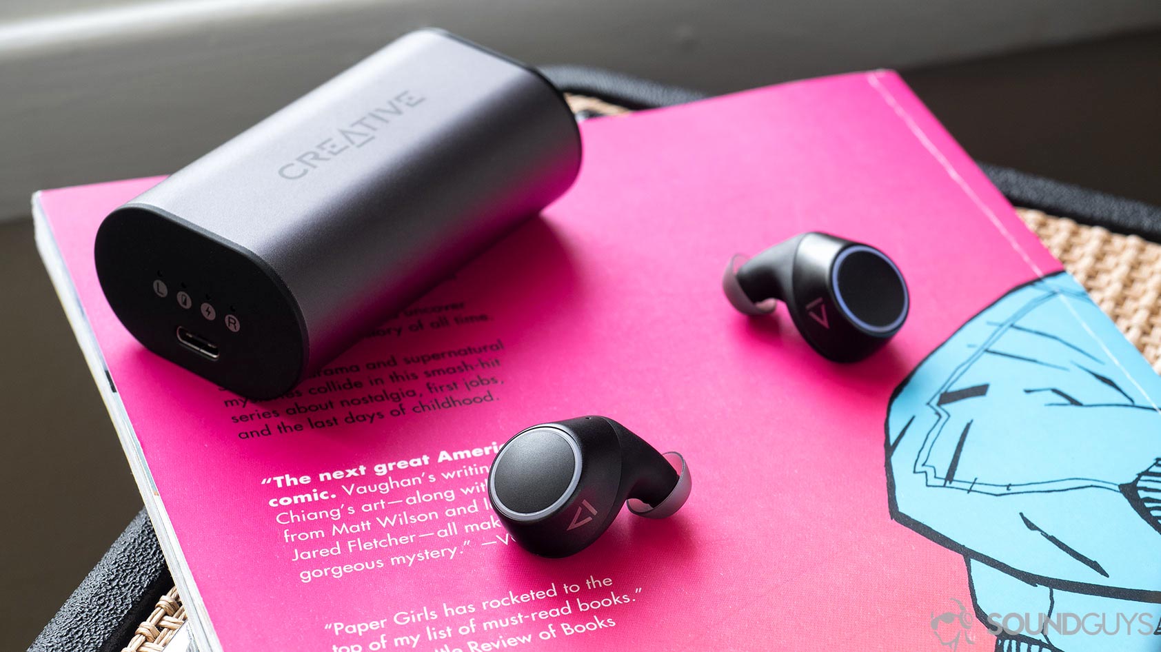 The Creative Outlier Air earbuds on a pink comic book with the charging case in the background.