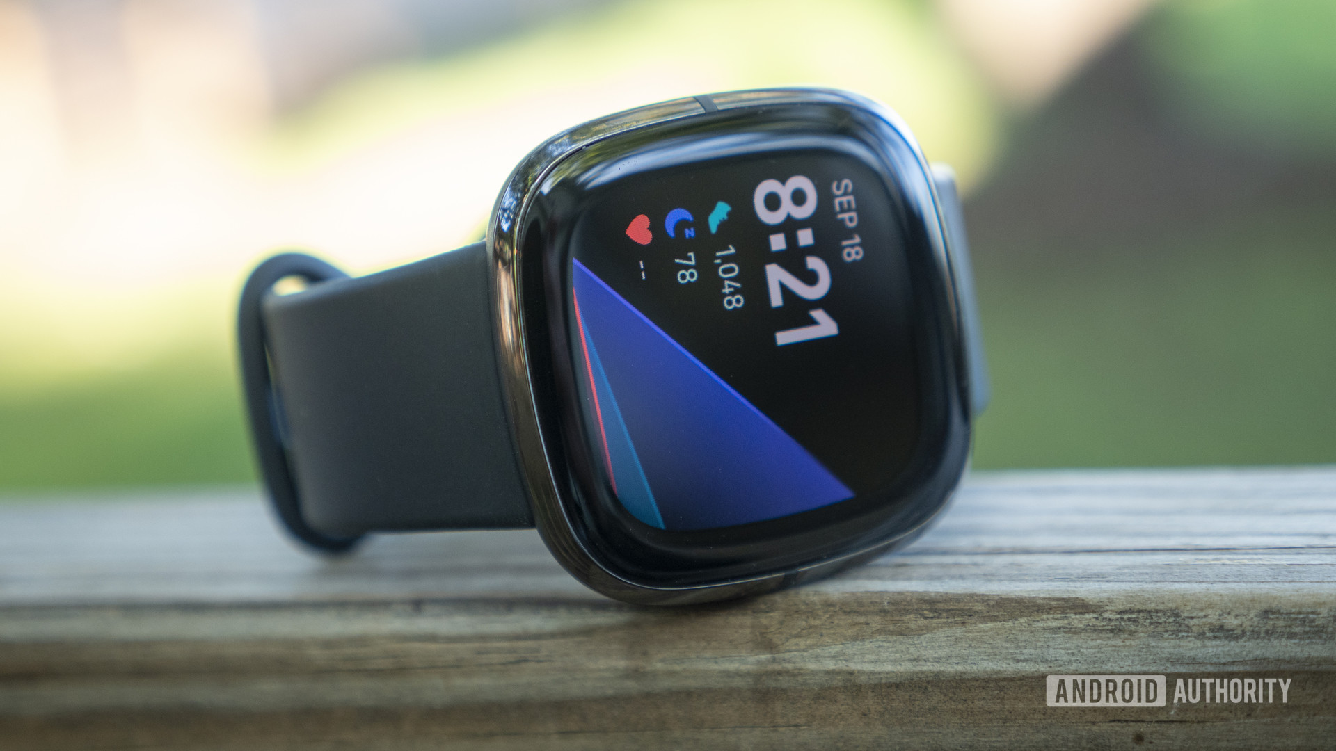 A Fitbit Sense rests on a wooden surface outdoors, displaying watch face 5.
