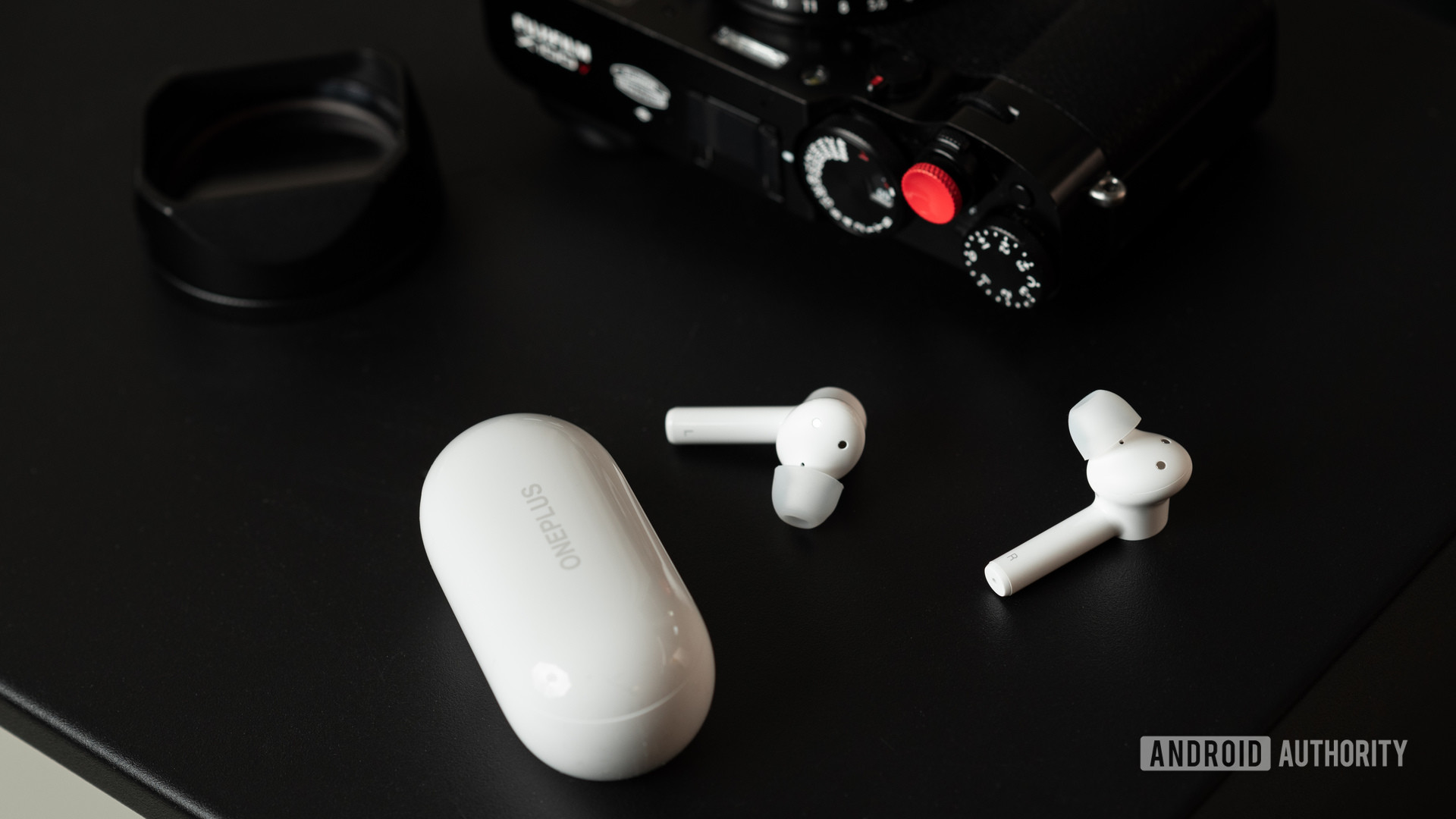 Les véritables écouteurs sans fil OnePlus Buds Z bon marché sur une table à côté d'un appareil photo et de l'étui de chargement.
