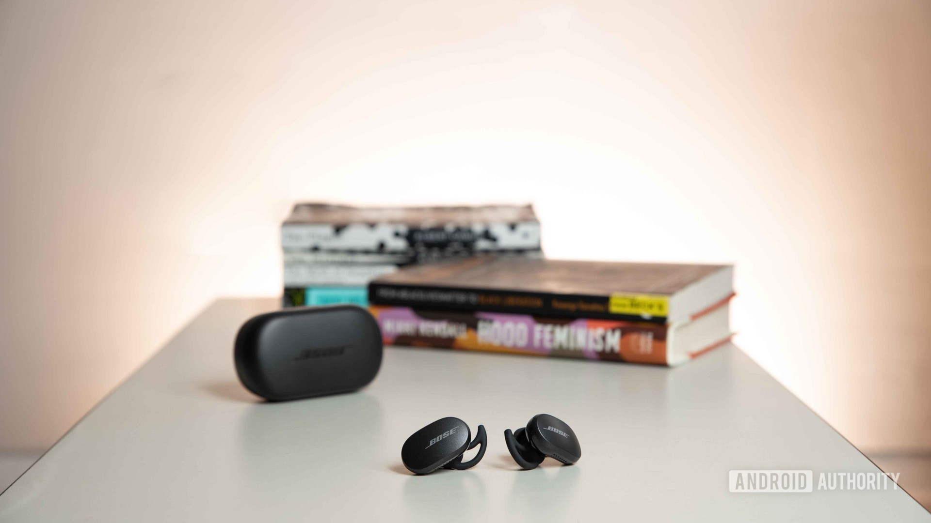 The Bose QuietComfort Earbuds noise cancelling true wireless earbuds rest outside of the charging case on a table and in front of a stack of books.