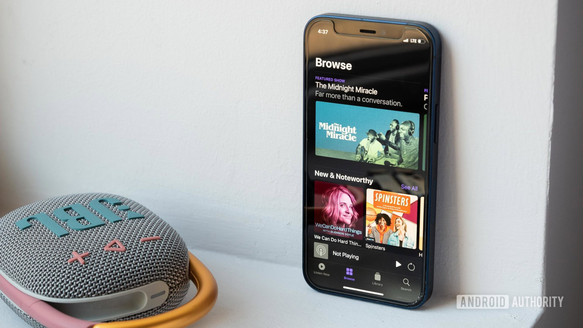 An iPhone 12 Mini rests against a wall, next to the JBL Clip 4 Bluetooth speaker, and the phone displays the Apple Podcast app homepage.