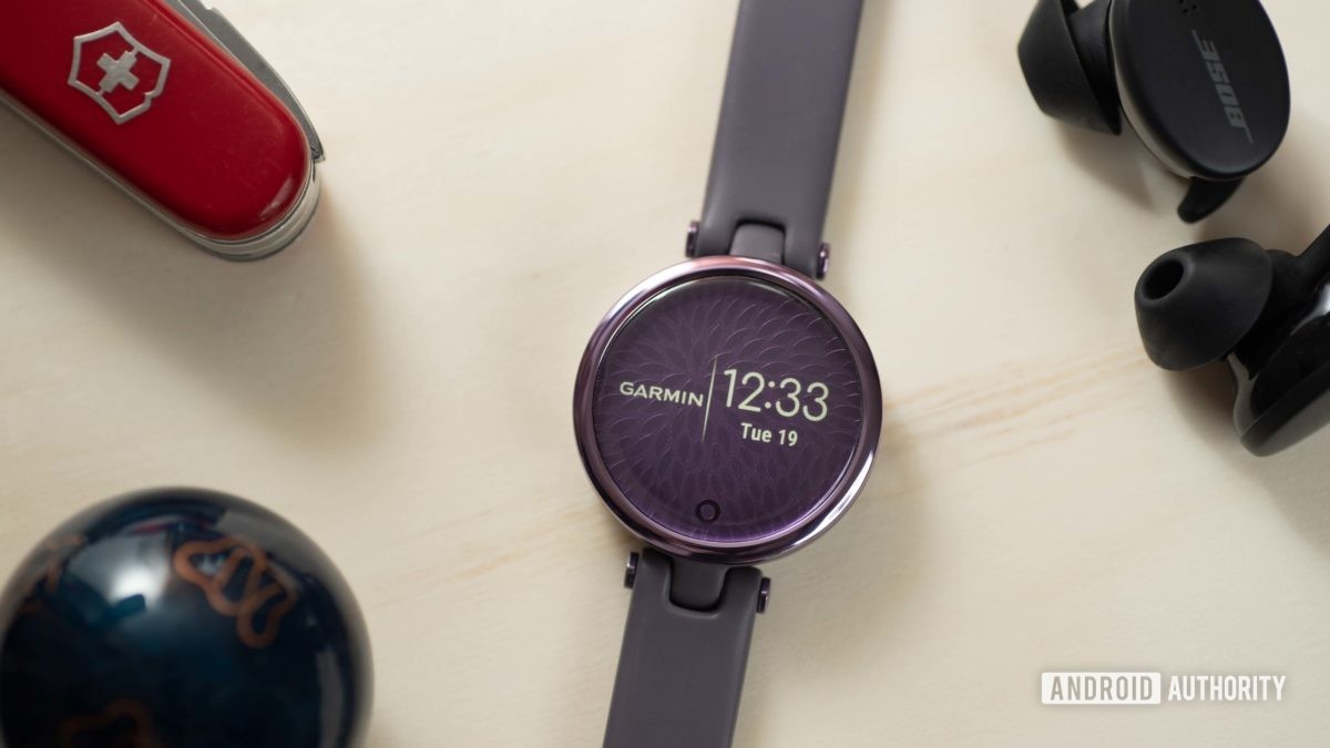 The Garmin Lily Sport Edition smart watch displays the time and Garmin logo, and rests next to a pair of Bose Sport Earbuds and a Swiss Army knife.