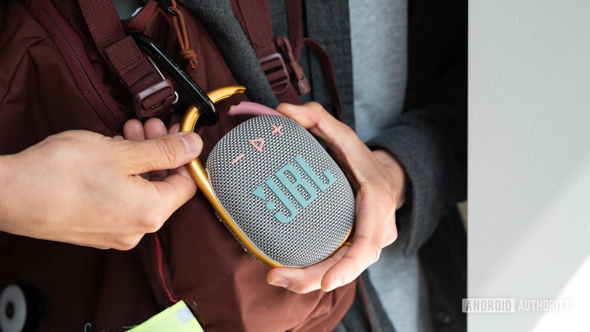 Two hands attach the JBL Clip 4 Bluetooth speaker to a red backpack.