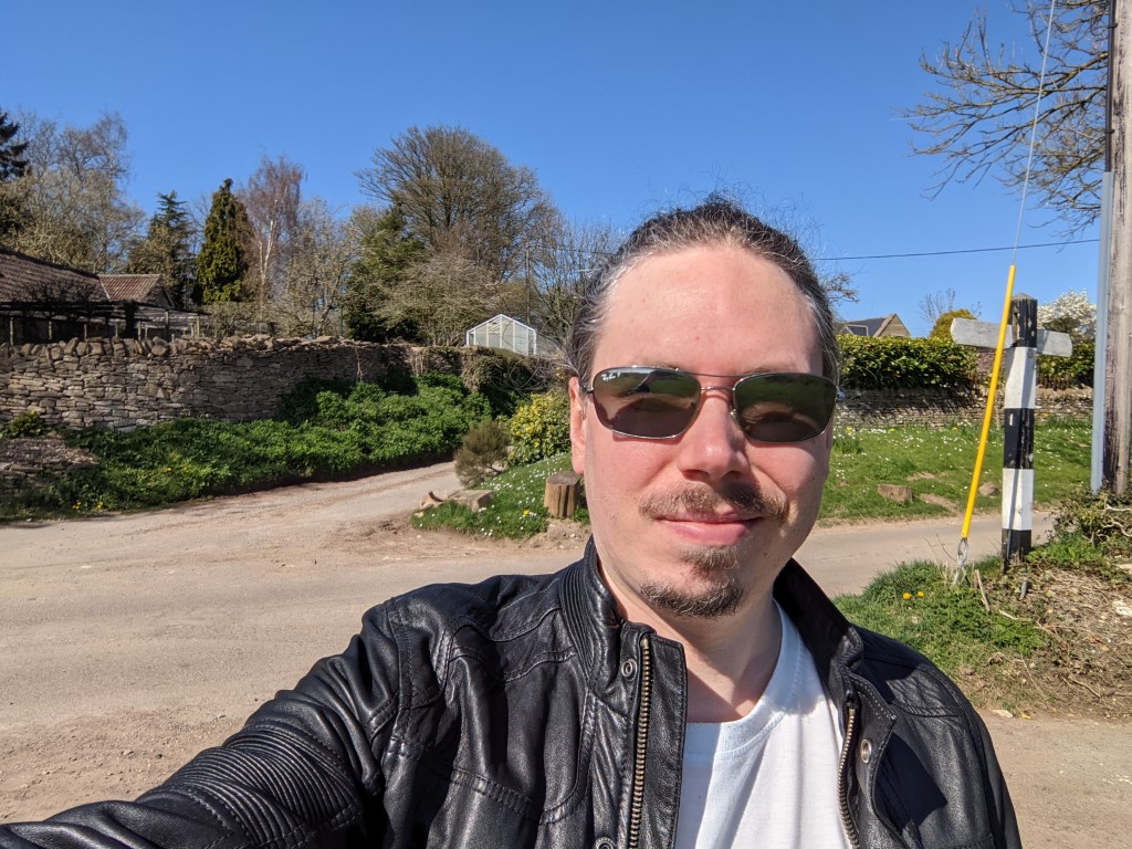Google Pixel 5 camera selfie 1 outdoor shot of a man with dark hair tied back and facial hair wearing shades and a leather jacket with a white t-shirt.