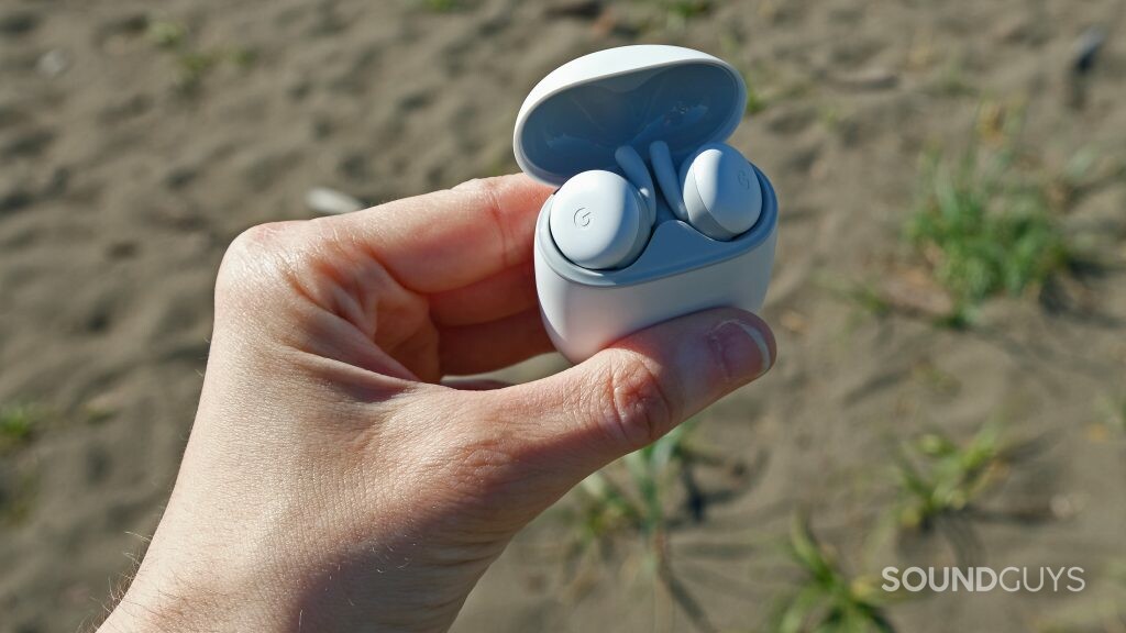 Une main tient le boîtier ouvert des Google Pixel Buds A-Series devant une plage.