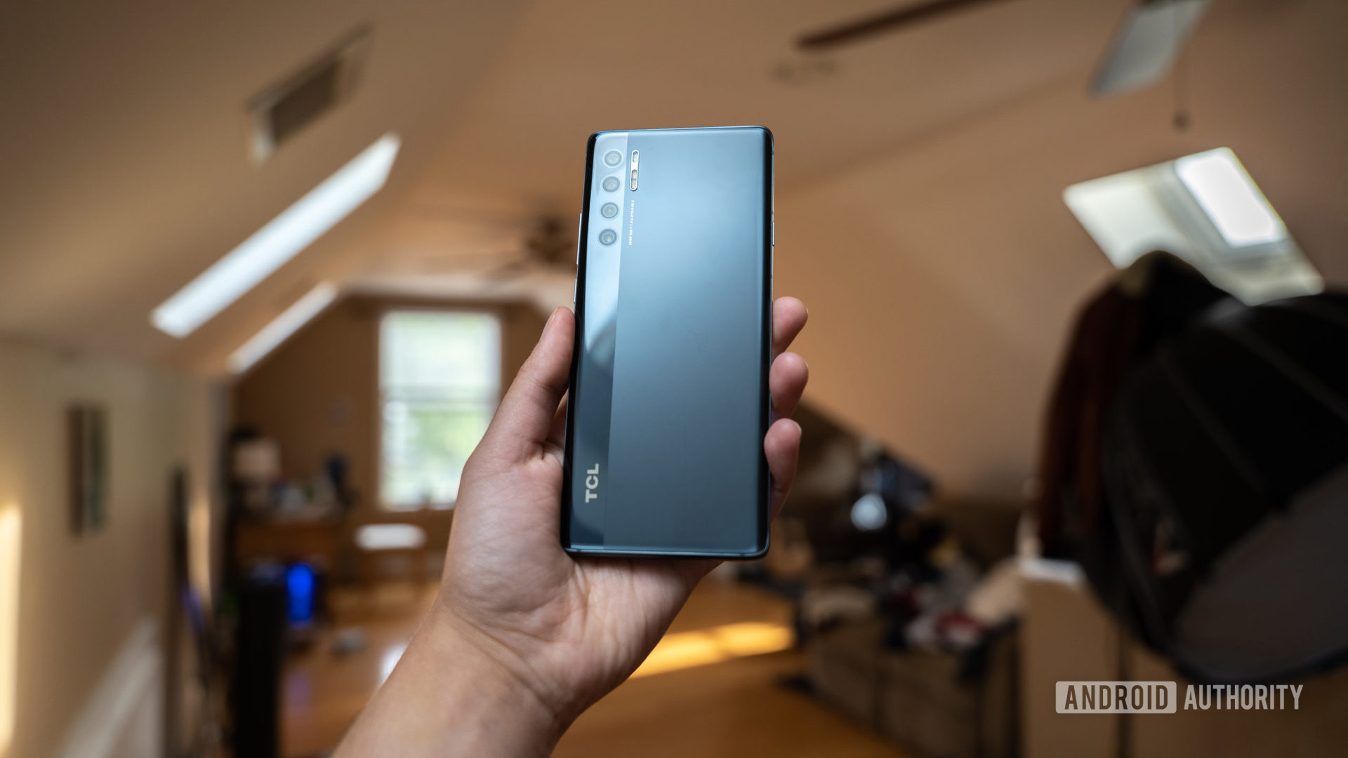 A hand holding up the TCL 20 Pro 5G showing the rear view of the phone indoors