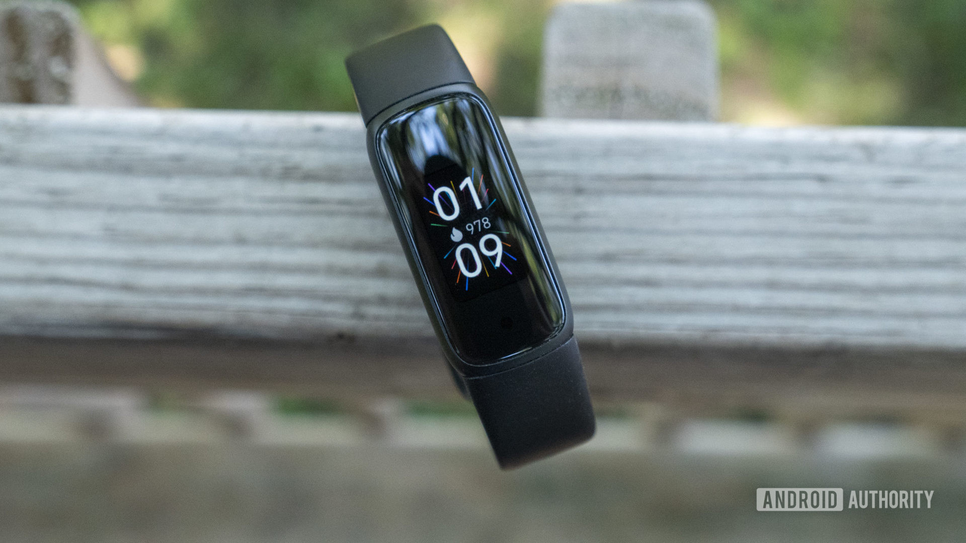 A Fitbit Luxe rests on a wooden fence with the watch face displayed.