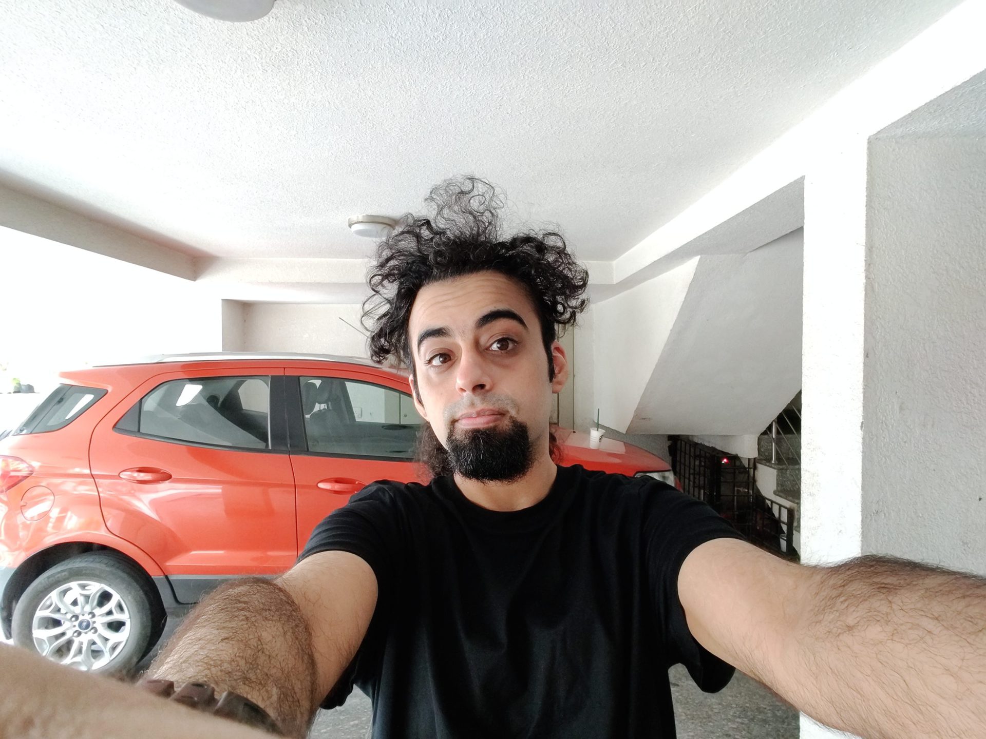 OnePlus Nord ultrawide selfie indoors of a man with black hair and a beard, wearing a black t-shirt and standing in front of a red vehicle.