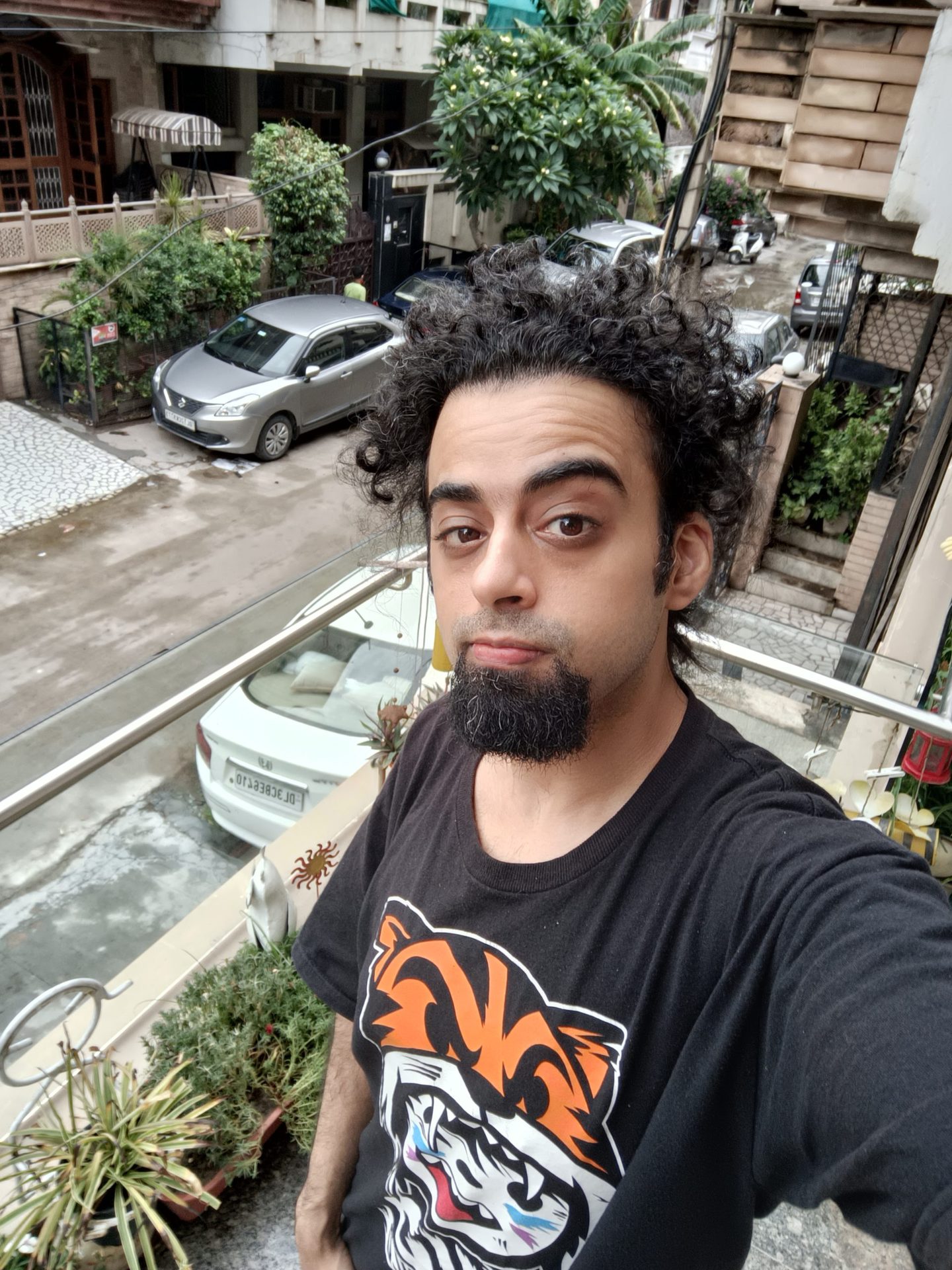 The Oppo Reno 6 Pro selfie standard showing a man with black hair and beard in a black t-shirt with orange pattern standing on a balcony above the street below.