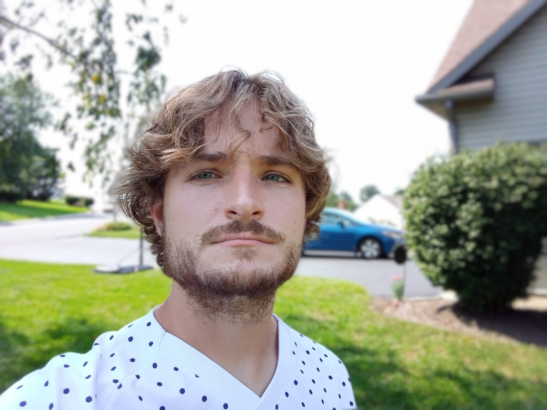 A portrait selfie of a man with light hair and facial hair wearing a white patterned t-shirt outdoors taken with the LG Premier Pro Plus 2