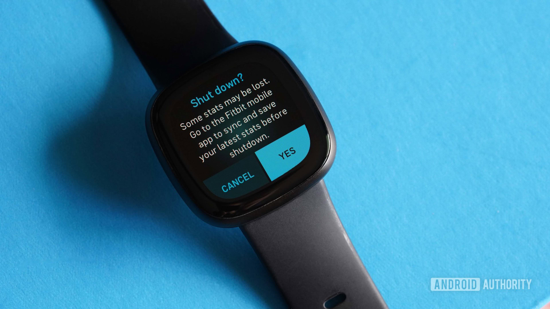 A Fitbit Versa 3 rests on a blue surface displaying the Shut Down confirmation screen.