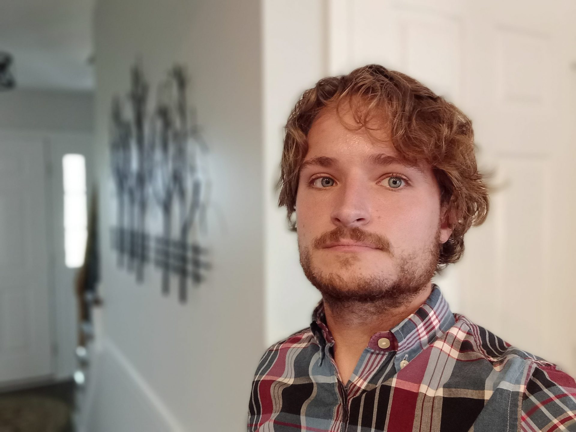 Galaxy a12 portrait selfie of a man with light hair and facial hair, wearing a red and black plaid shirt, taken indoors.