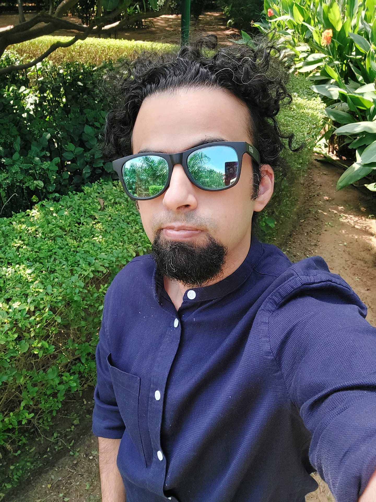 Samsung Galaxy A52s 5G selfie camera standard shot of man with dark hair and beard wearing a blue long-sleeve shirt and mirrored shades, taken outdoors next to a green hedge.