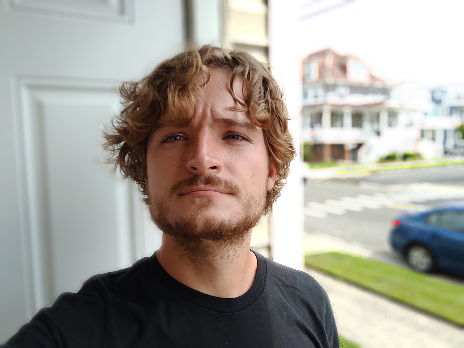 A portrait selfie taken with the ZTE Blade X1 of a man with light curly hair and a beard, wearing a black t-shirt and standing in an open doorway.