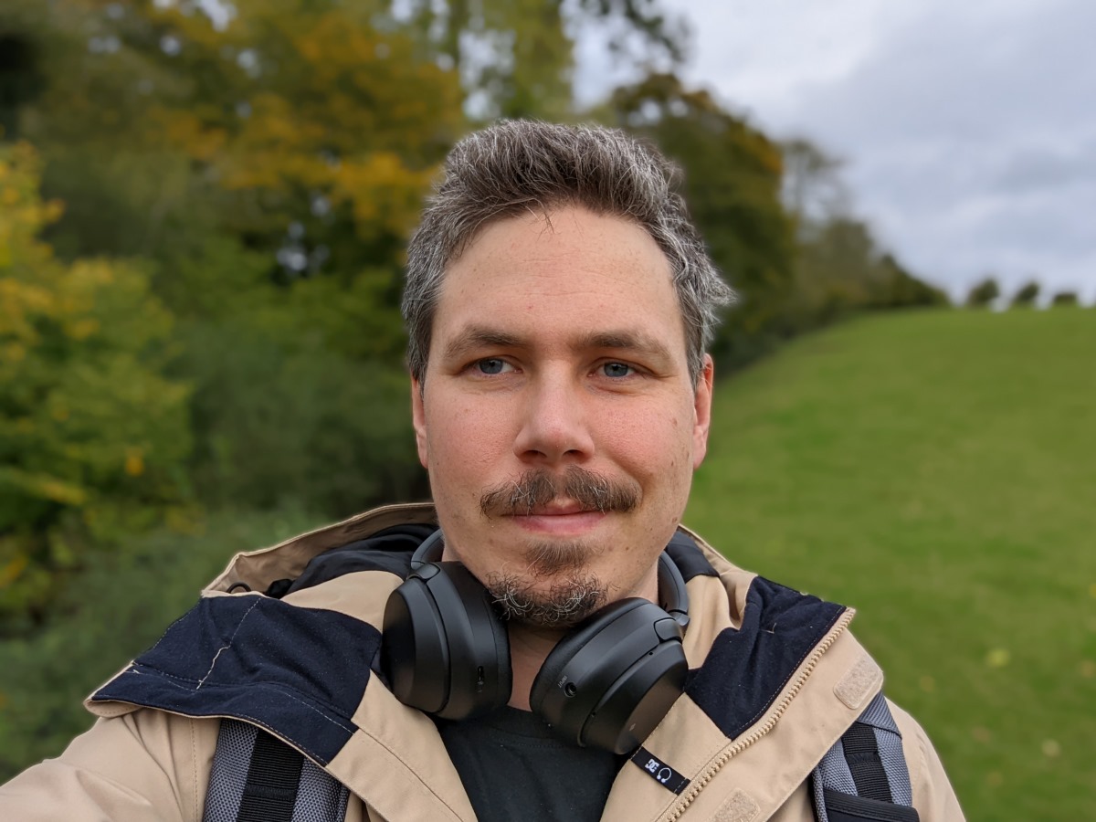 Camera sample selfie outdoor portrait shot of a man with dark hair and facial hair wearing a beige jacket, with headphones around his neck, taken on the Google Pixel 6 Pro