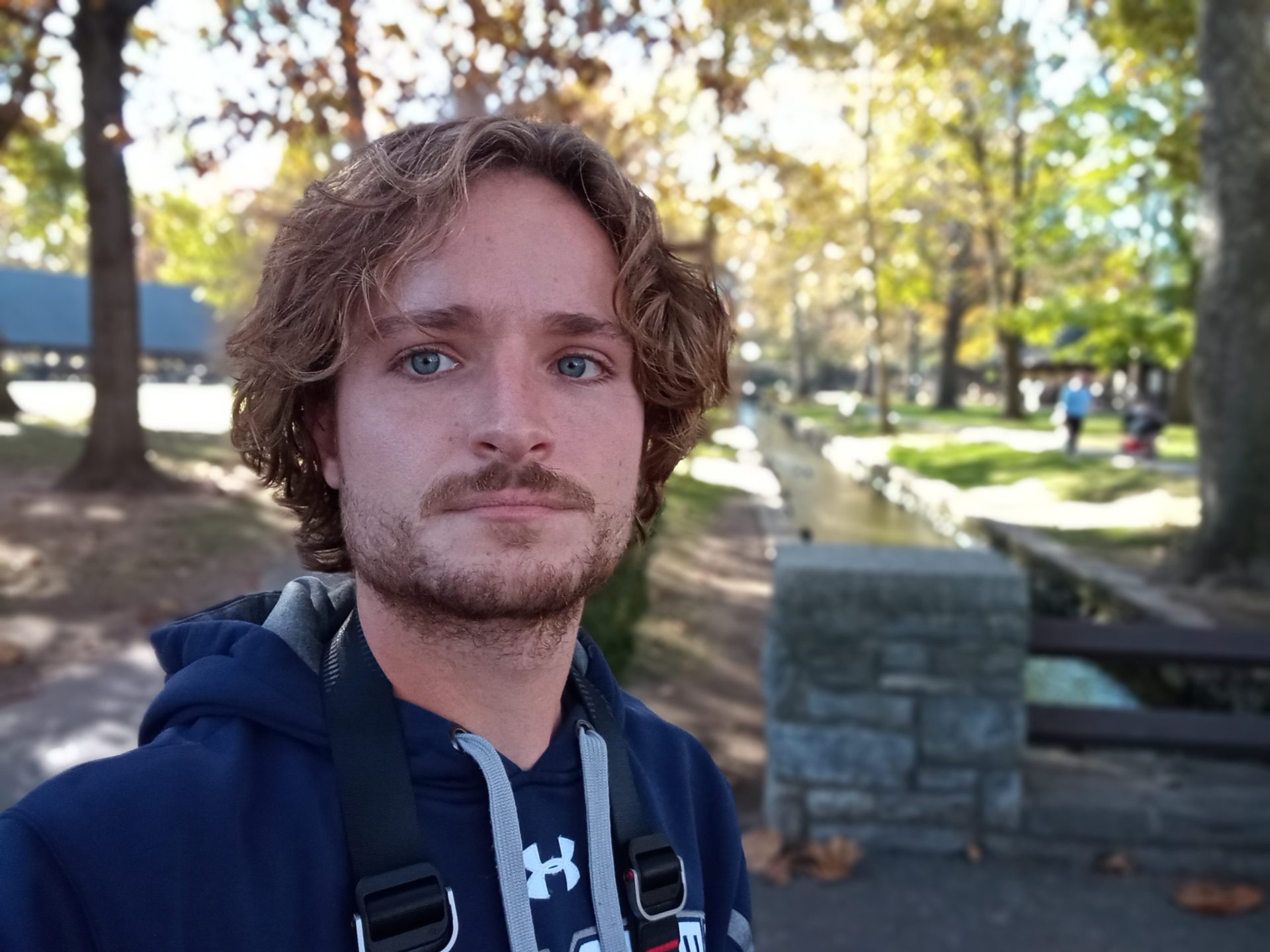 moto g pure portrait selfie of a man with light curly hair and facial hair, wearing a t-shirt and hoodie, with trees behind him.