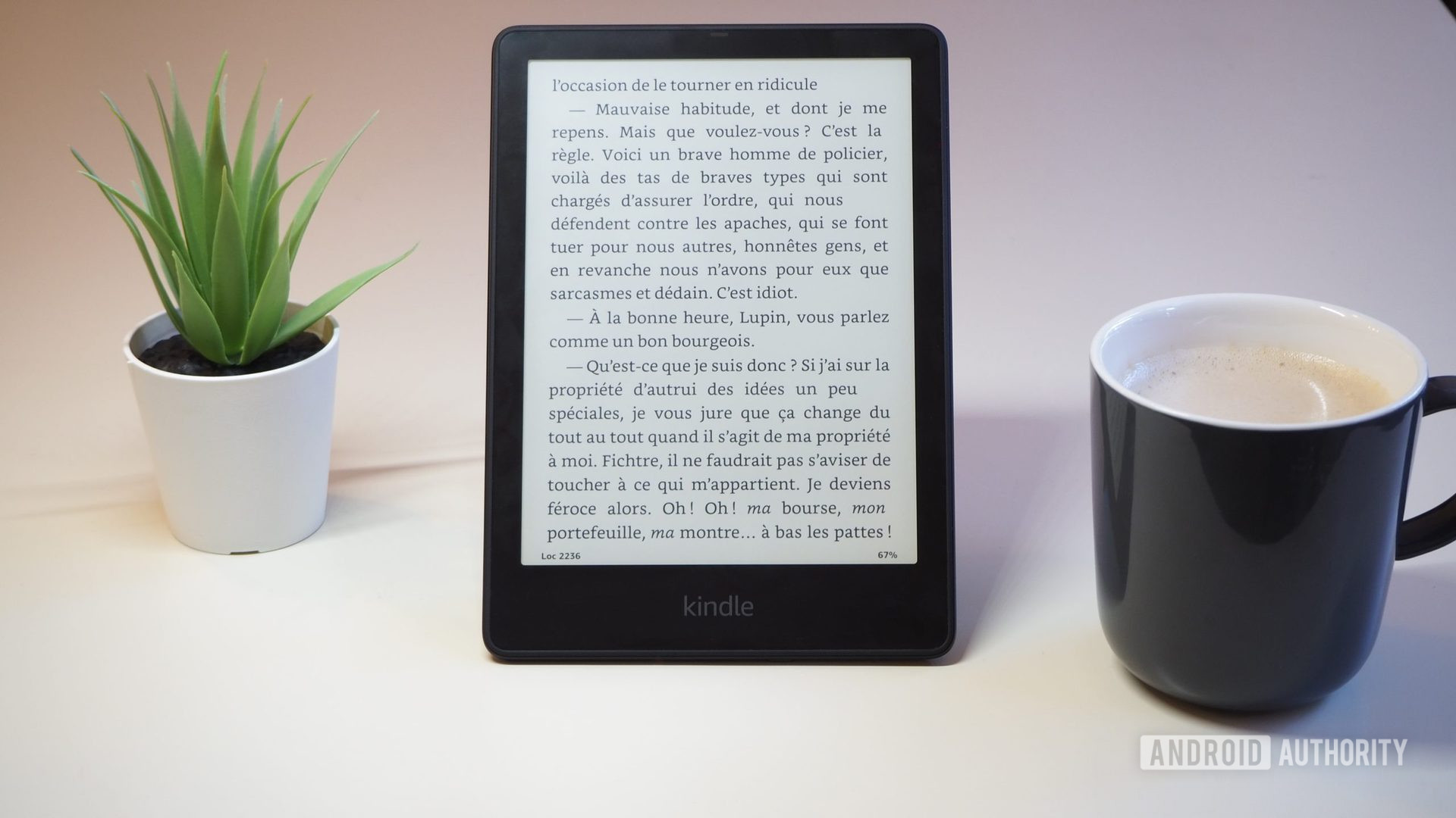 Amazon Kindle Paperwhite 2021 with the screen showing a book, on a table, next to a cup of coffee and plant, version 1
