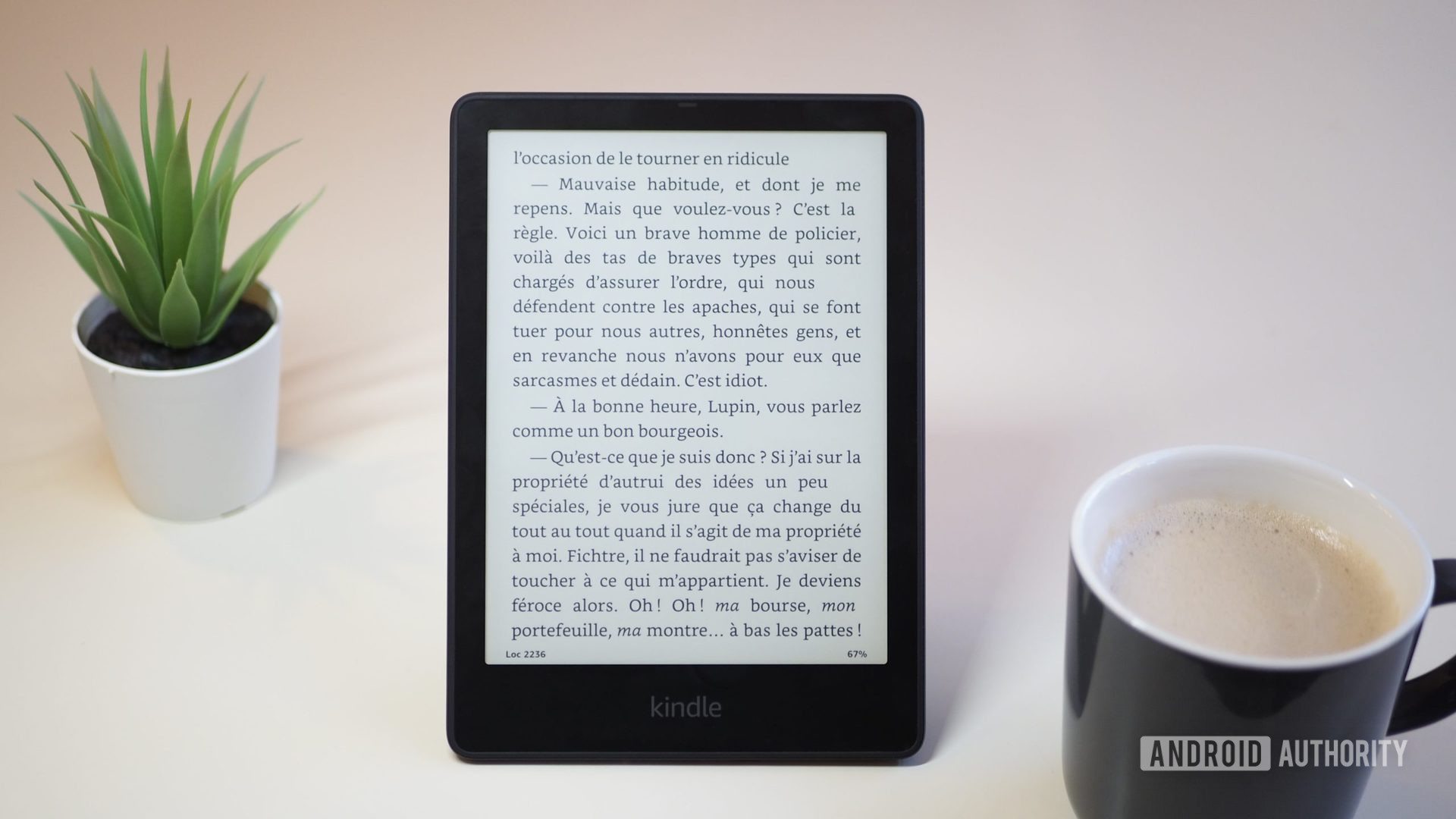 Amazon Kindle Paperwhite 2021 with the screen showing a book, on a table, next to a cup of coffee and plant, version 2