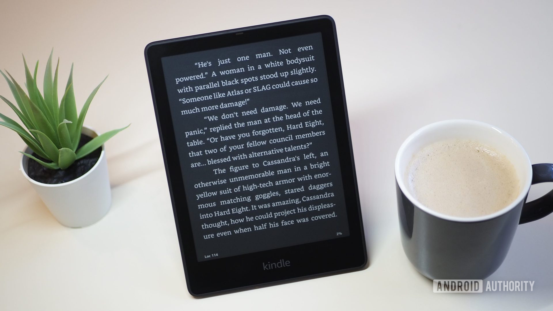 Amazon Kindle Paperwhite 2021 with the screen in dark mode showing a book, on a table, next to a cup of coffee and plant