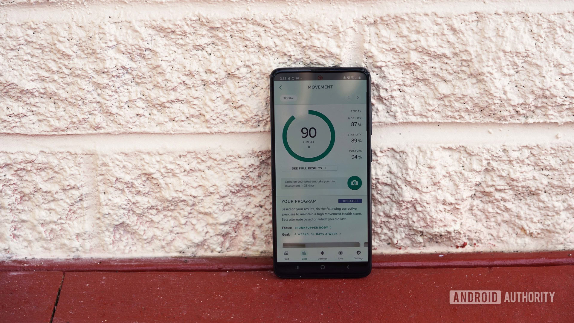 A Samsung Galaxy A51 rests against a white brick wall displaying a user's Movement Health score in the Amazon Halo app.