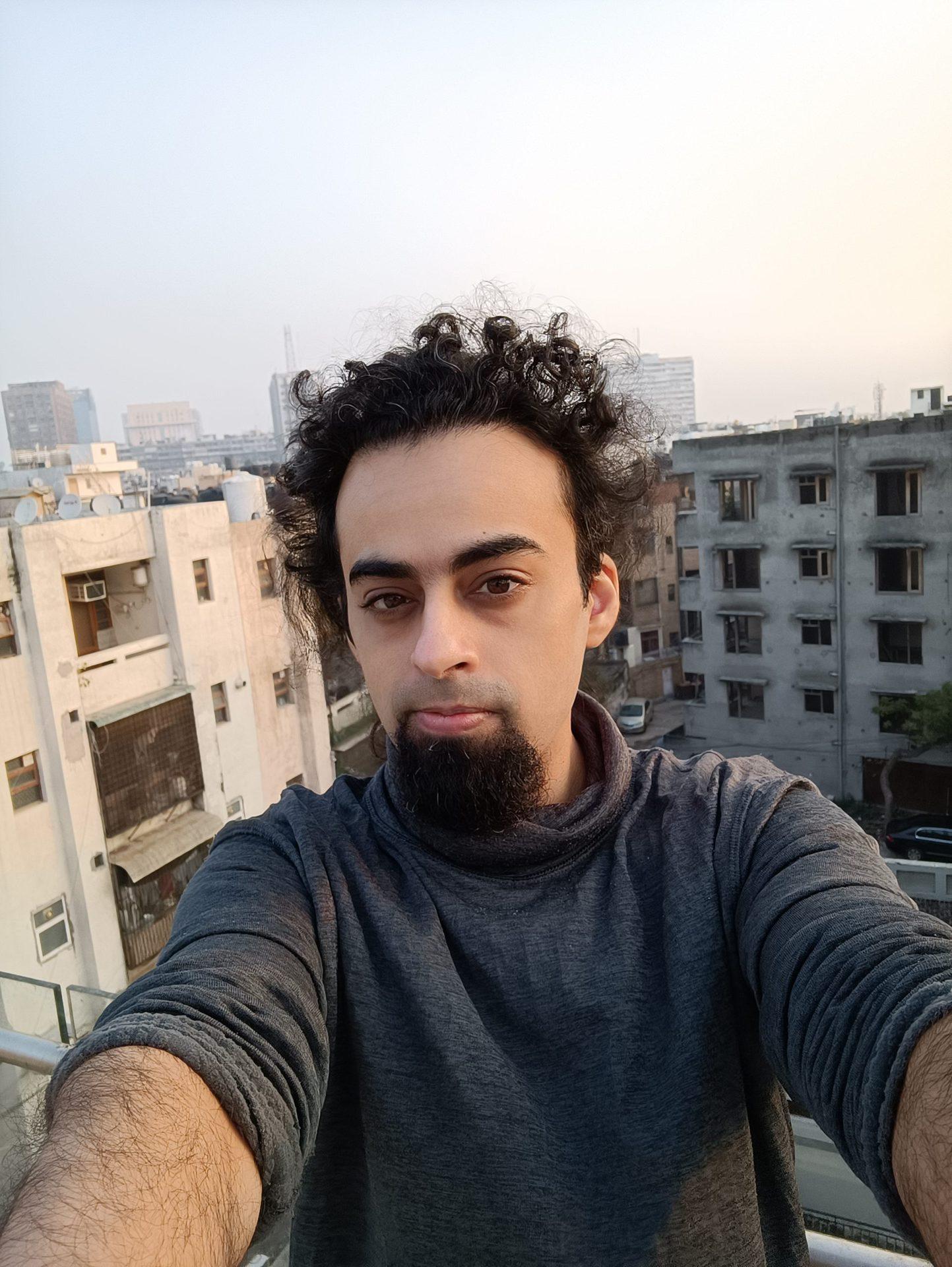 Mi 11i câmera frontal selfie de um homem com cabelo escuro encaracolado e barba, com edifícios visíveis atrás dele