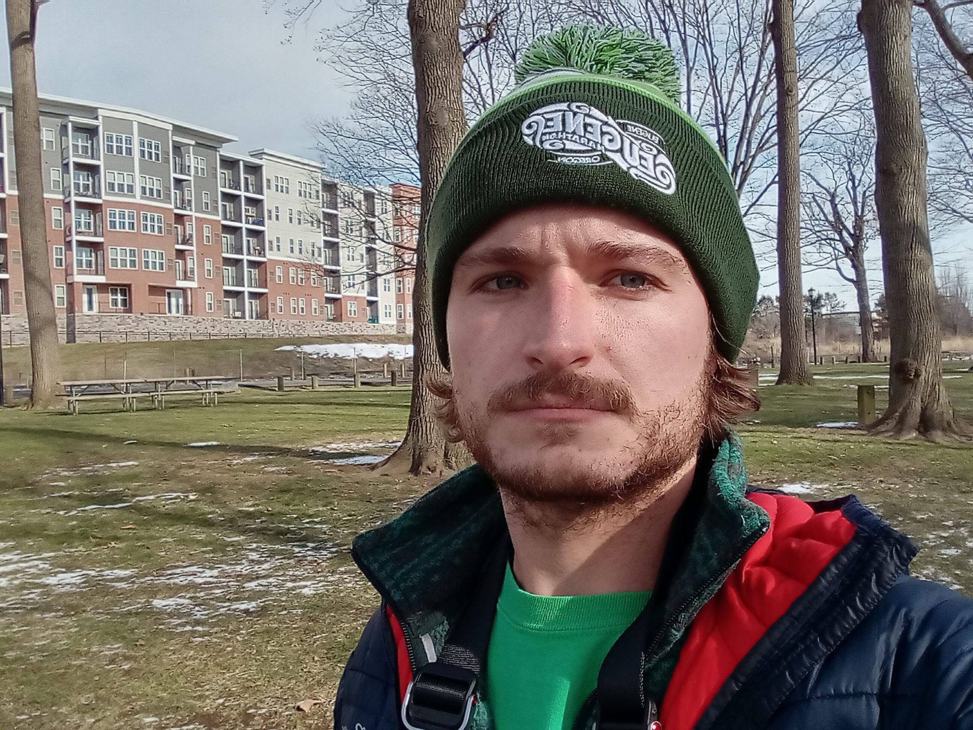 galaxy a03s standard selfie outdoors of a man with facial hair wearing a green hat, green t-shirt, and red and black coat, with buildings and trees visible behind him.